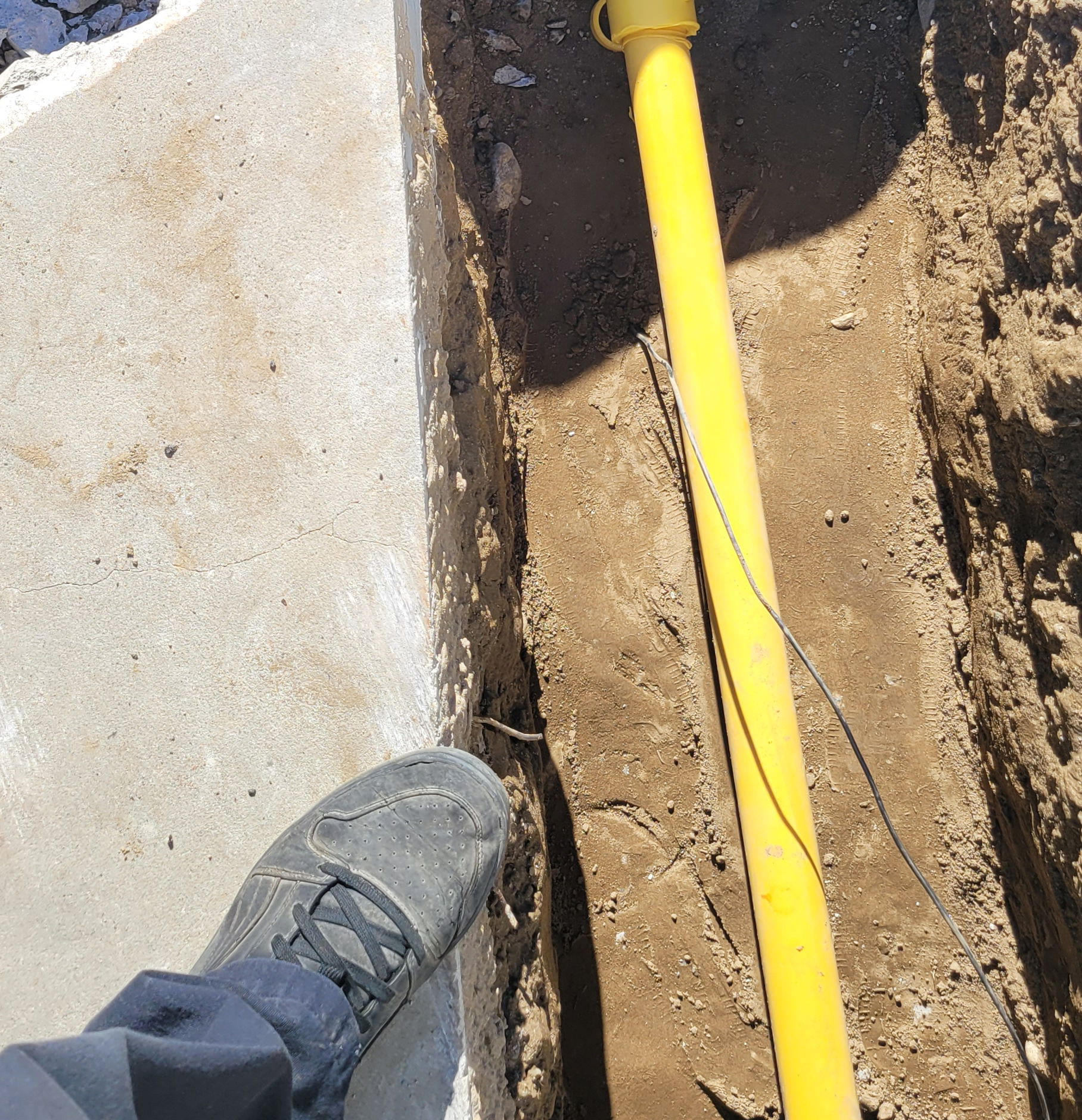 A plumber wearing black pants and shoes stands near a trench containing a yellow pipe and a thin black wire beside a concrete surface.
