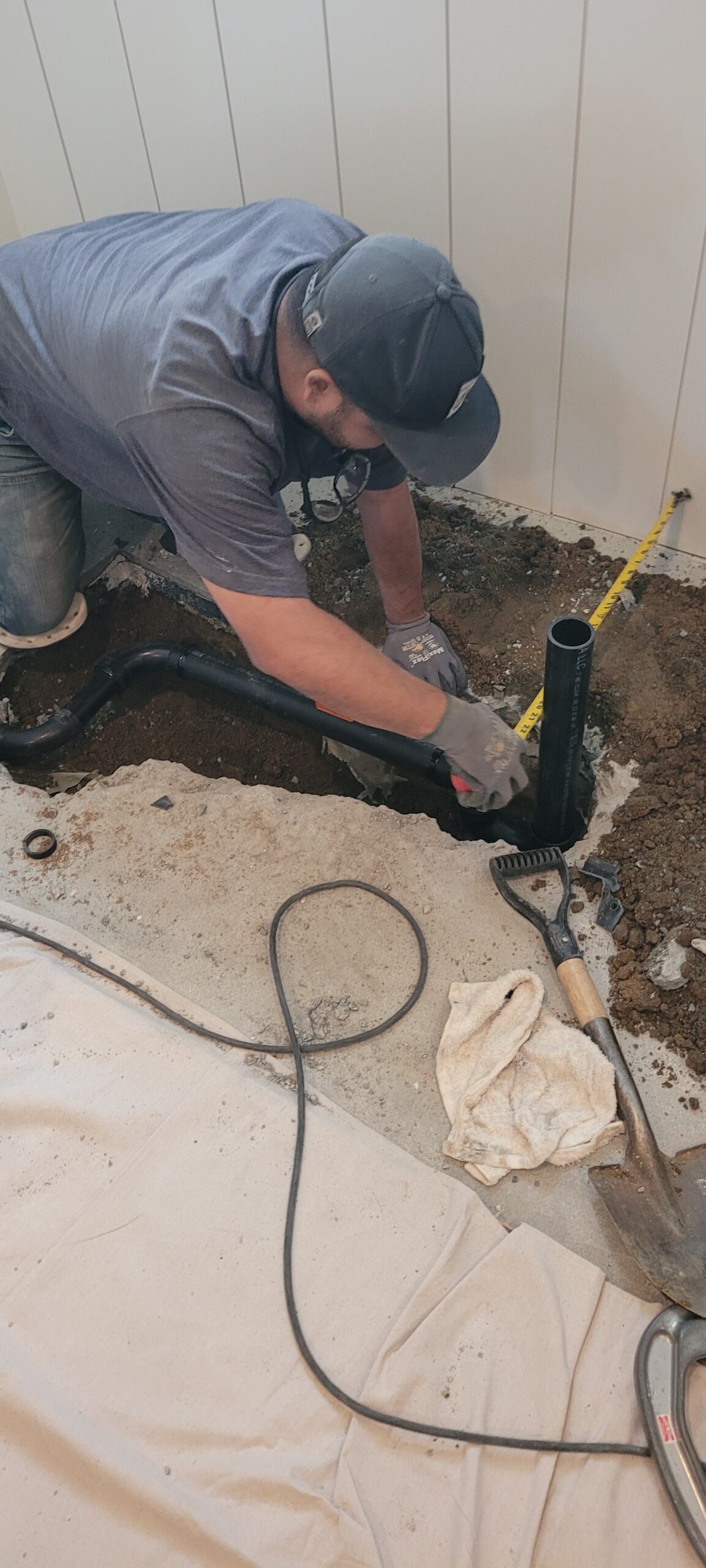 A person measures and works on exposed pipes in a floor trench, surrounded by tools, dirt, and a measuring tape, during a plumbing installation or repair inside a building.