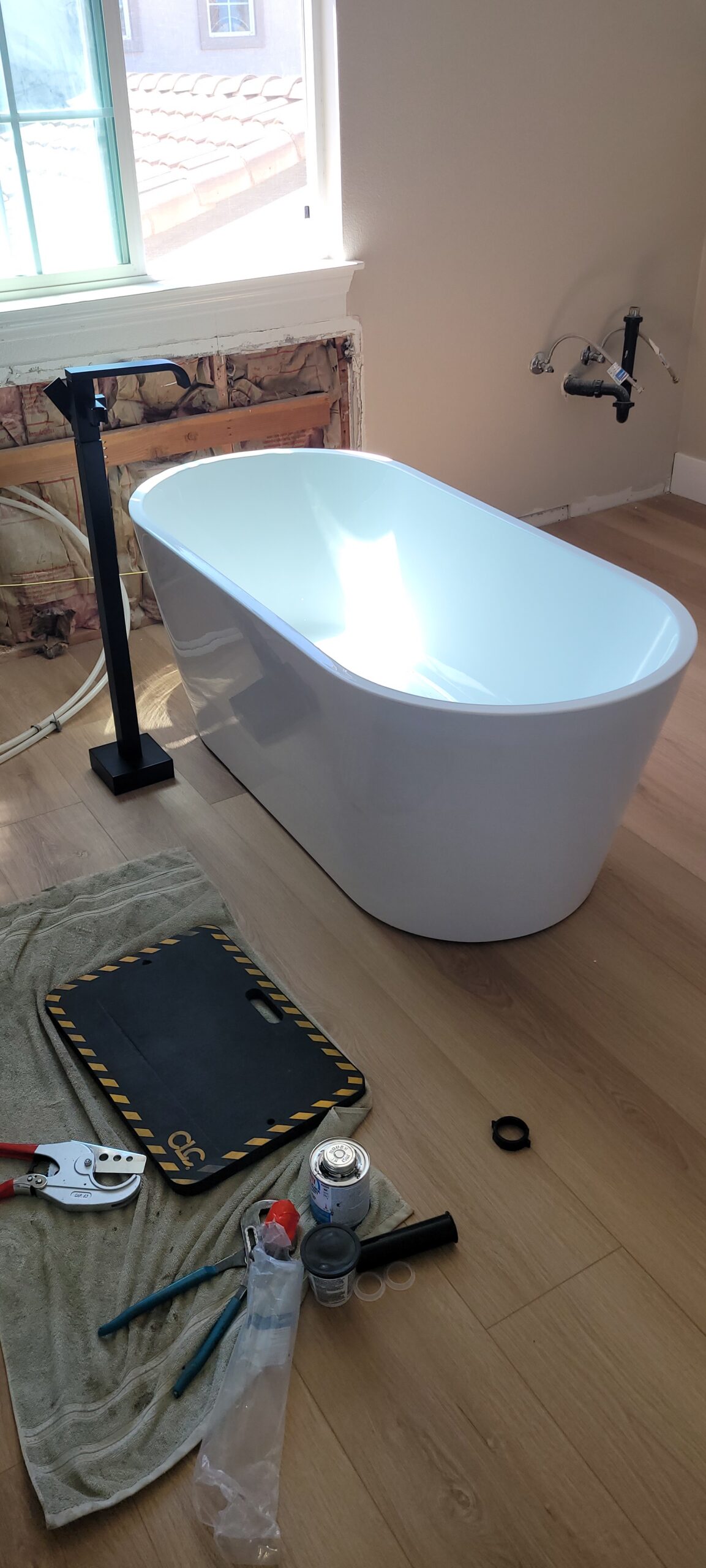 A freestanding bathtub is being installed in a bathroom; plumbing tools, pipes, and parts are scattered on the floor nearby. The wall behind the tub is unfinished.