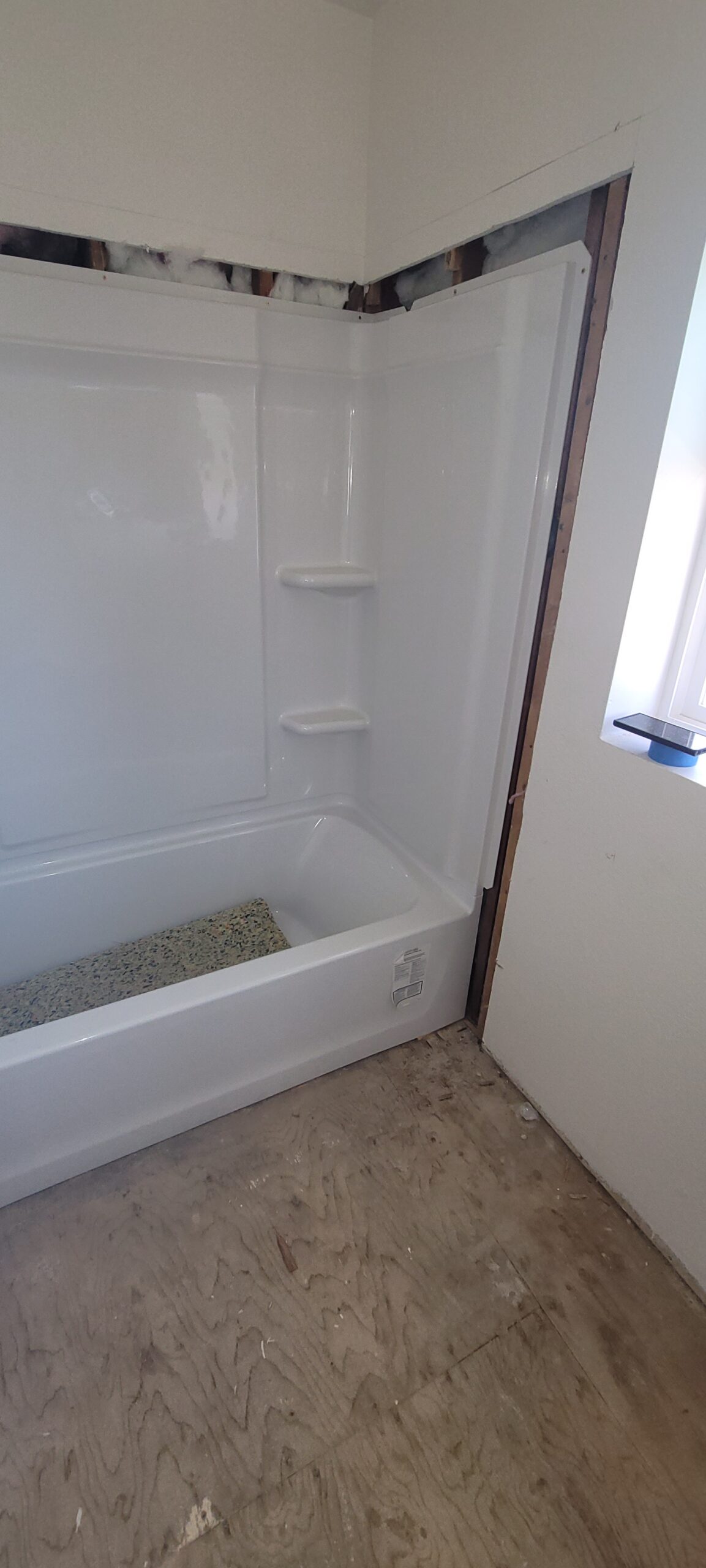 A bathroom under renovation with an installed bathtub and exposed wall framing above and beside the tub; subfloor is visible.