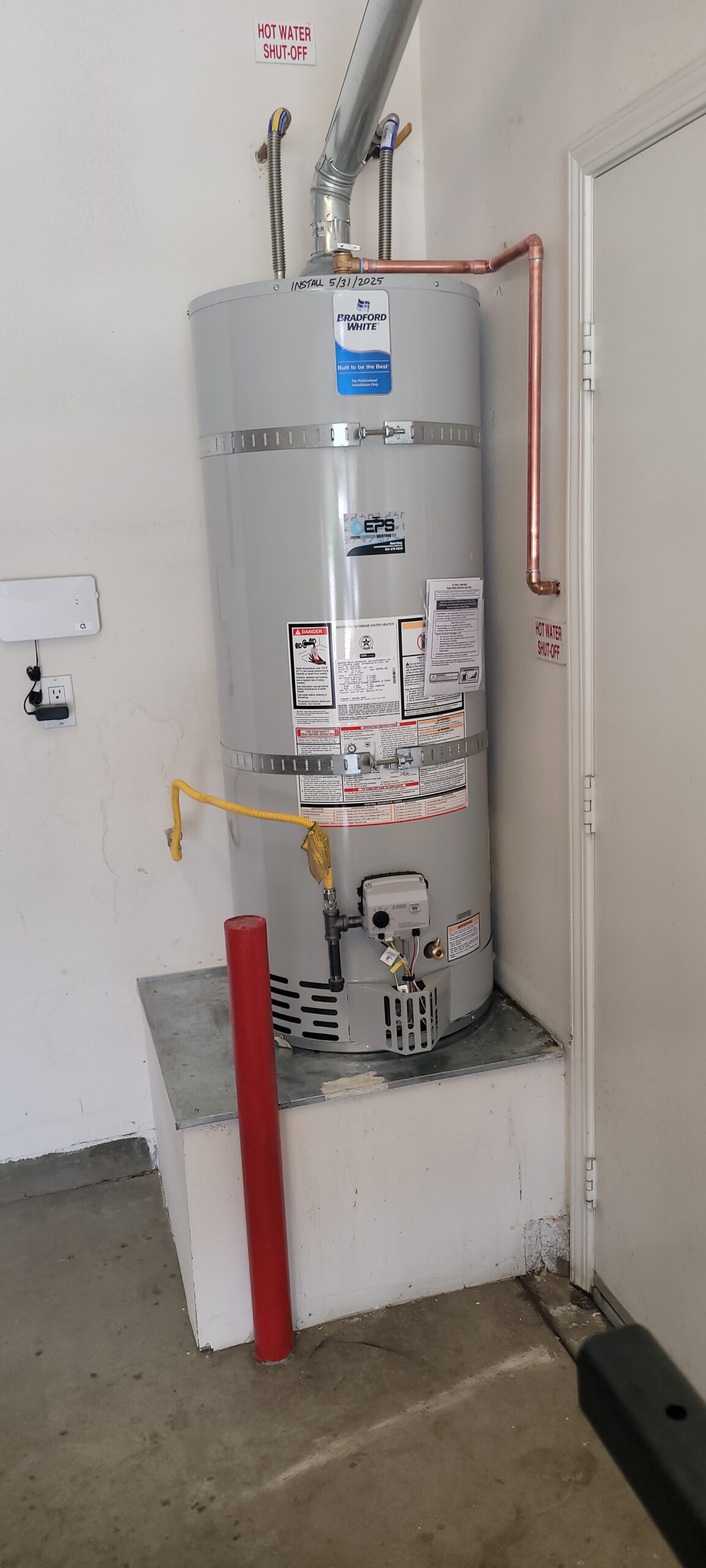 A gray water heater is installed on a raised platform in a garage, with pipes, warning labels, and a red safety post nearby.