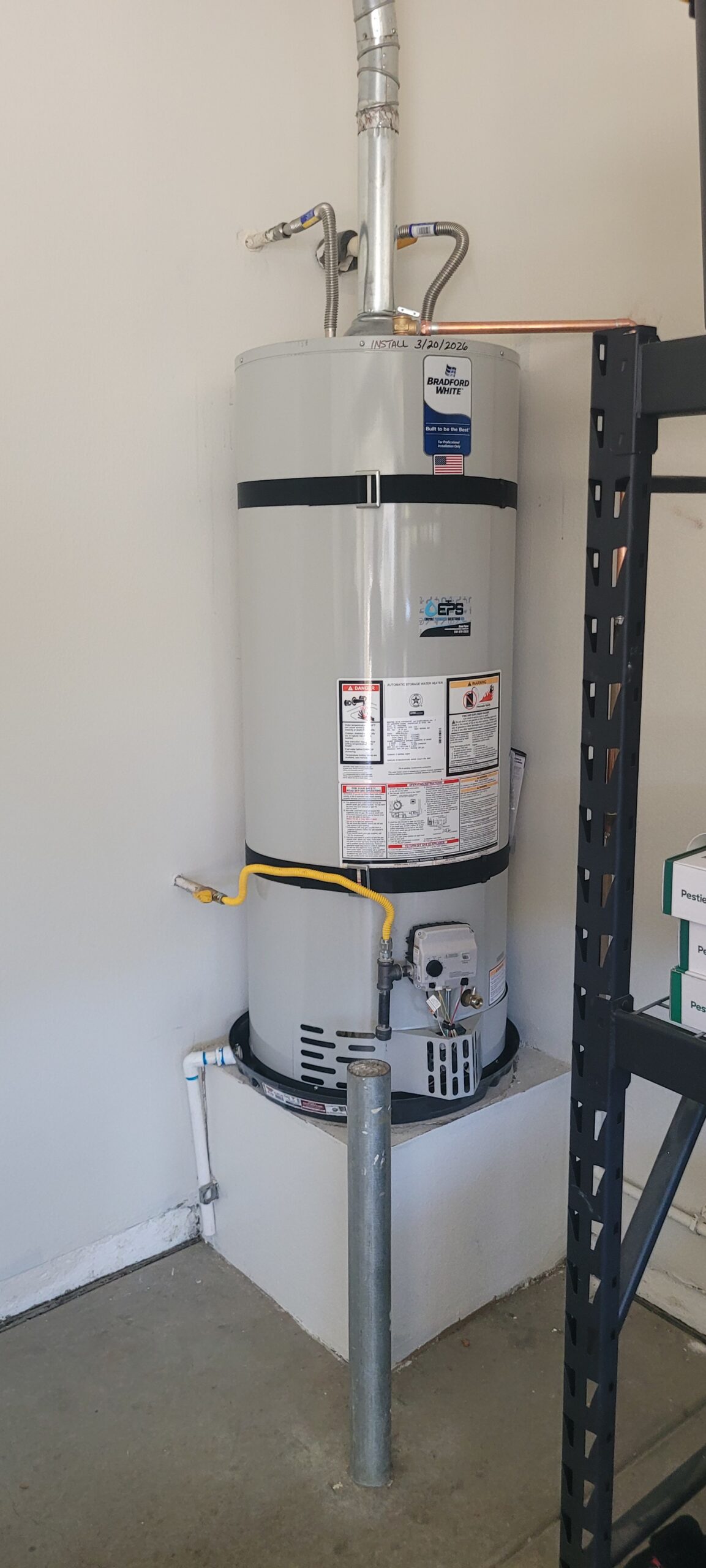 A gray water heater is installed in a corner of a garage, with yellow and silver pipes attached and safety labels visible on the front.