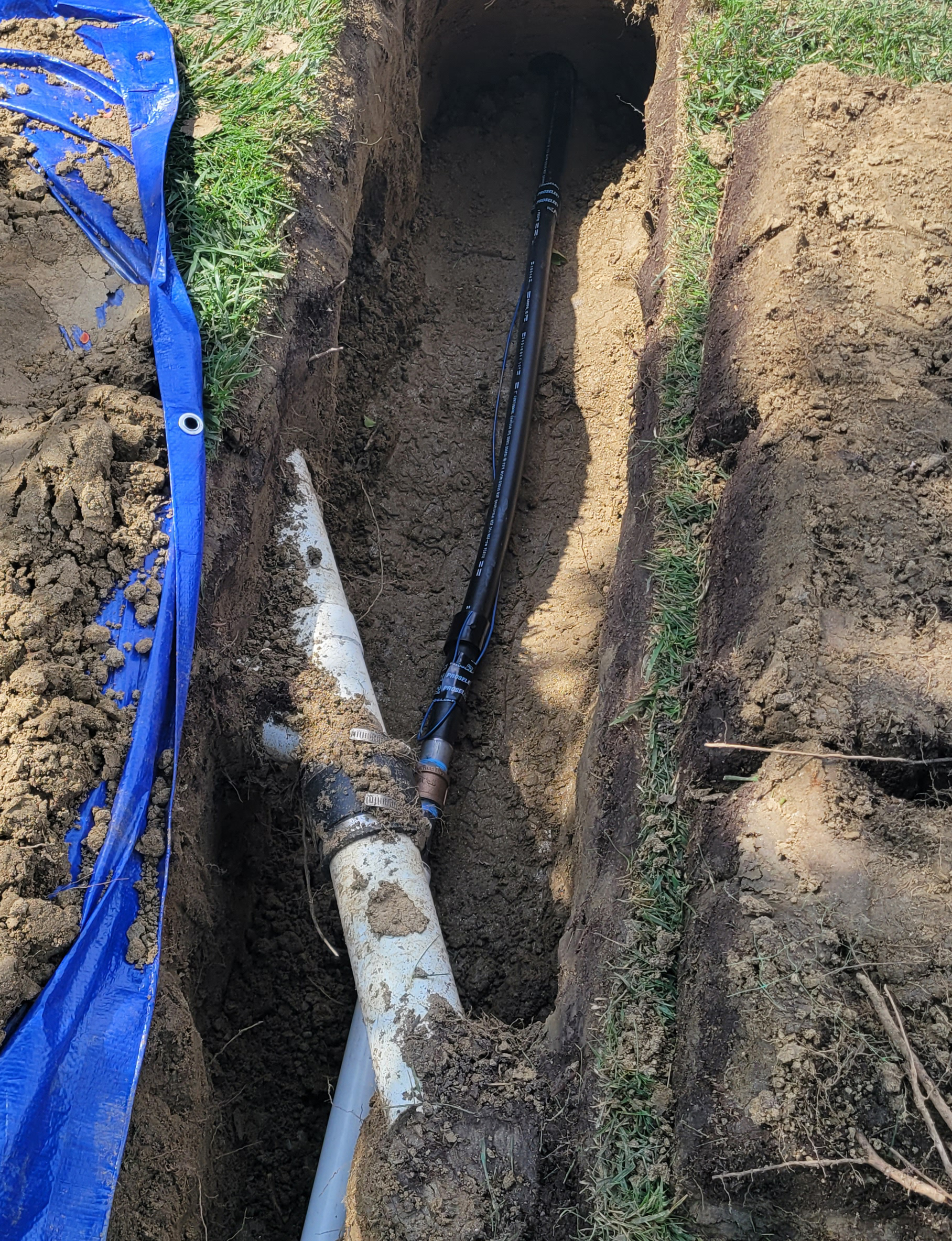 A trench in the ground, likely prepared by a skilled plumber, shows exposed pipes, including a white PVC pipe and a black conduit, partially covered with soil and surrounded by grass.