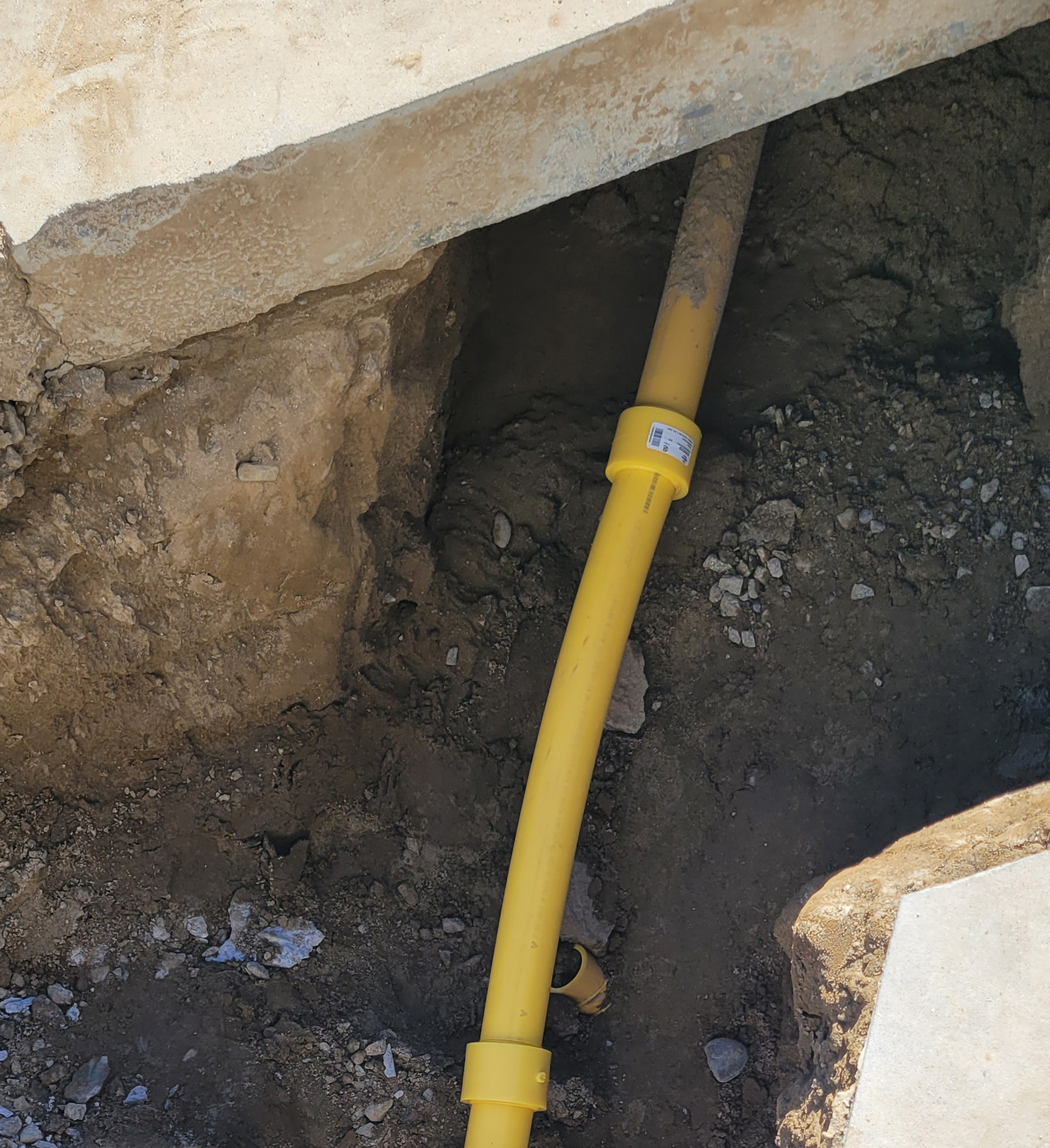 Yellow plastic pipe runs through a dirt trench under a cut concrete slab, possibly indicating utility installation or repair work by a plumber.