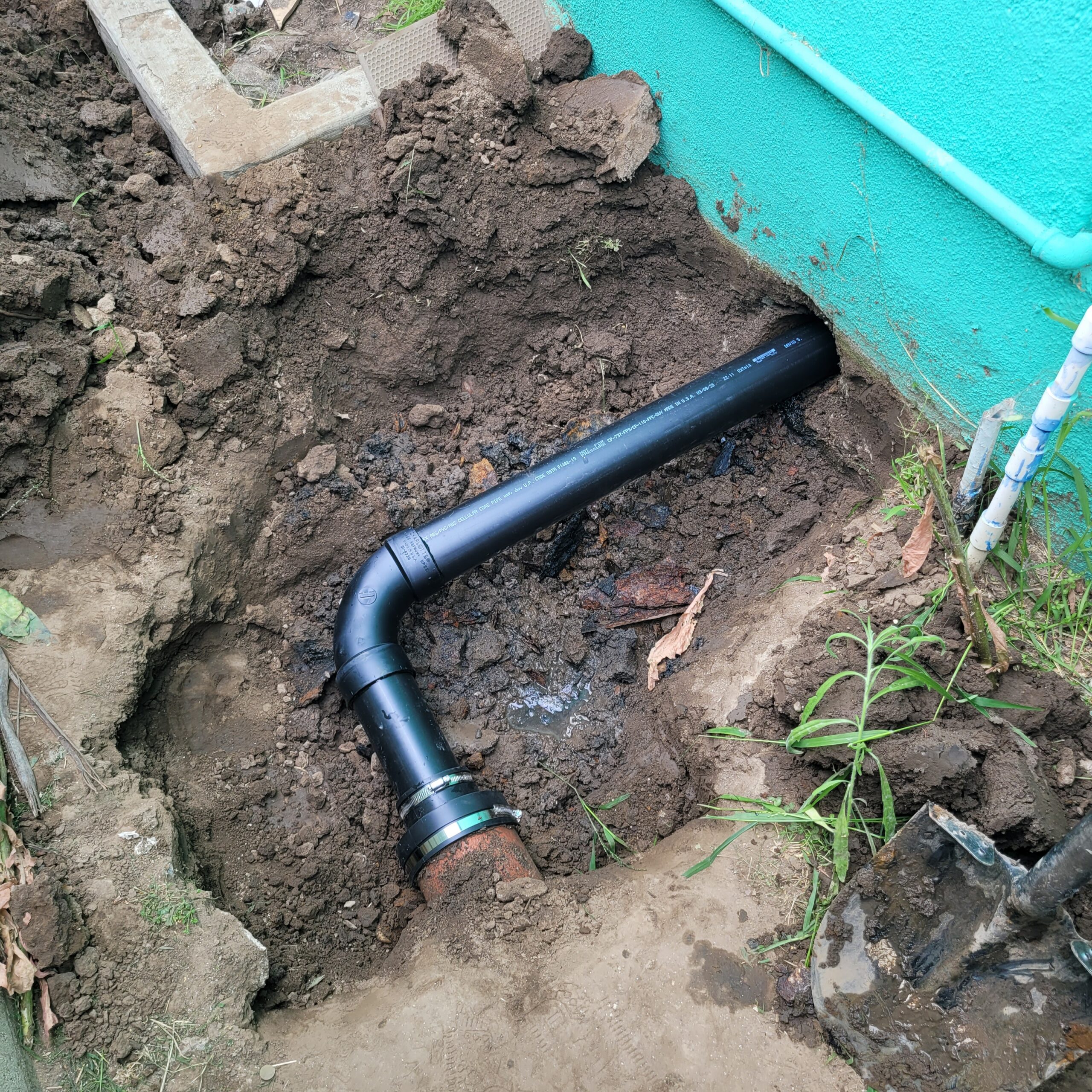 A partially buried black PVC drainage pipe with an elbow joint is connected to a turquoise wall, surrounded by soil and construction tools, indicating recent work by a skilled plumber.
