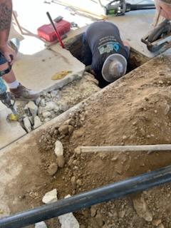 A worker is digging into a hole in a concrete floor, surrounded by tools, dirt, and rubble, as part of a sewer line repair and replacement project, while another person stands nearby.