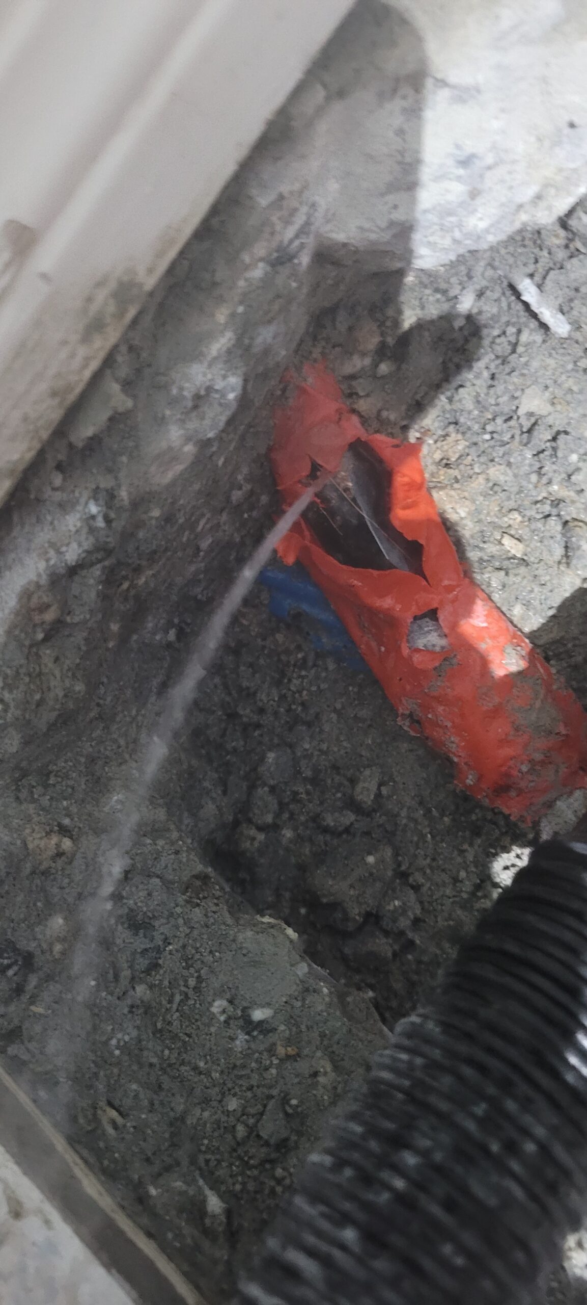 A stream of water is leaking from a damaged pipe wrapped in red tape, surrounded by dirt and concrete debris, signaling the urgent need for a plumber.