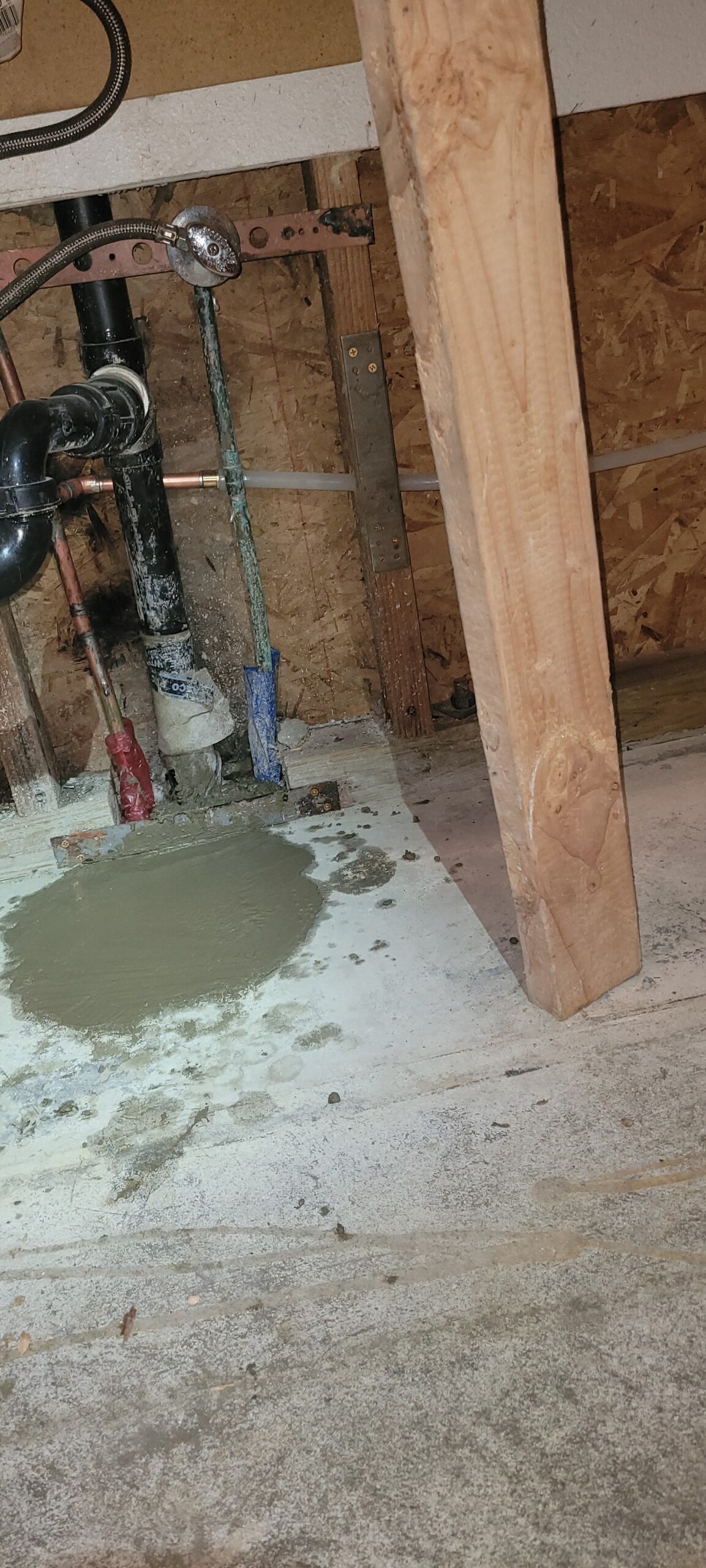 Exposed plumbing pipes, likely installed by a skilled plumber, and a fresh concrete patch on the floor next to a wooden support post inside an unfinished wall.